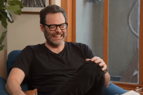 A Man Wearing Glasses And A Black Shirt Is Sitting In A Chair Laughing GIF