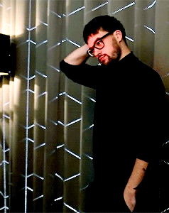 A Man Wearing Glasses And A Black Shirt Is Standing In Front Of A Metal Wall GIF