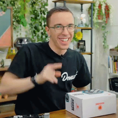 A Man Wearing Glasses And A Black Shirt With The Word Donut On It Is Sitting At A Table With A Box On It . GIF