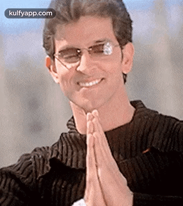 A Man Wearing Glasses And A Black Sweater Is Smiling And Praying With His Hands Folded . GIF