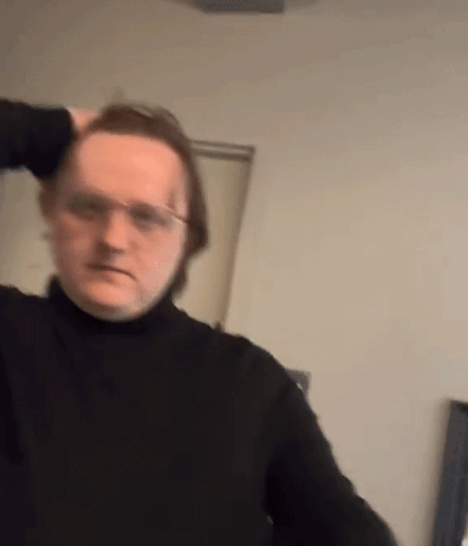 A Man Wearing Glasses And A Black Turtleneck Is Holding His Hair . GIF