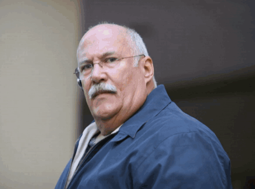 A Man Wearing Glasses And A Blue Jacket Looks At The Camera GIF