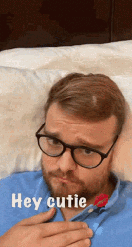 A Man Wearing Glasses And A Blue Shirt Is Laying On A Bed With The Words Hey Cutie Above Him GIF