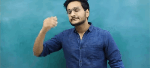 A Man Wearing Glasses And A Blue Shirt Is Standing In Front Of A Blue Wall And Making A Funny Face . GIF