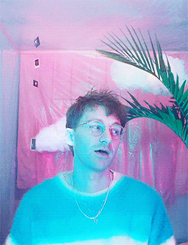 A Man Wearing Glasses And A Blue Shirt Stands In Front Of A Pink Curtain GIF