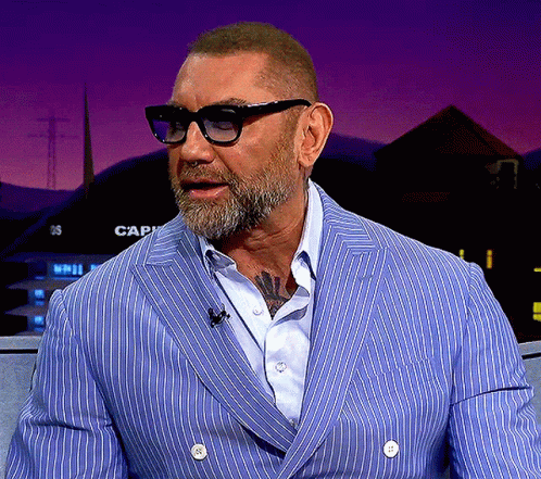 A Man Wearing Glasses And A Blue Striped Jacket Is Sitting In Front Of A Sign That Says Cap GIF