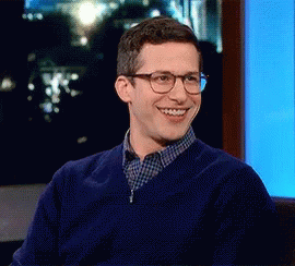 a man wearing glasses and a blue sweater is smiling while sitting in front of a screen .