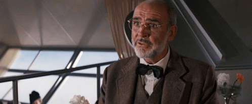A Man Wearing Glasses And A Bow Tie Is Sitting In Front Of A Window GIF
