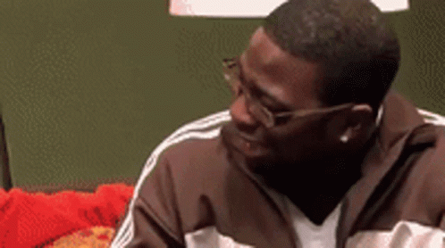 A Man Wearing Glasses And A Brown Jacket Is Sitting On A Red Couch . GIF