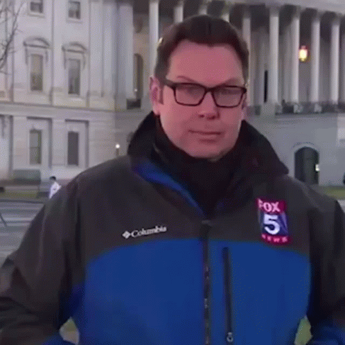 A Man Wearing Glasses And A Columbia Jacket Is Standing In Front Of A Large Building . GIF