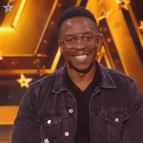 A Man Wearing Glasses And A Denim Jacket Smiles For The Camera GIF
