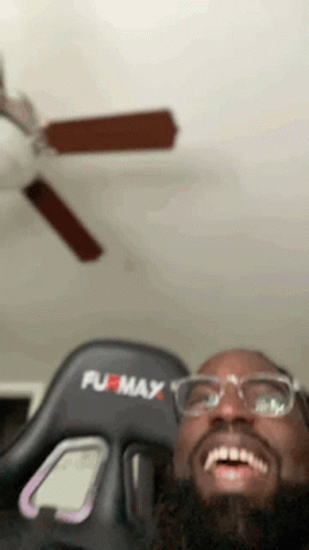 A Man Wearing Glasses And A Furmax Gaming Chair Is Smiling In Front Of A Ceiling Fan . GIF
