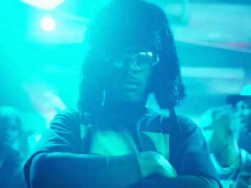 A Man Wearing Glasses And A Furry Hat Is Standing In Front Of A Crowd Of People GIF