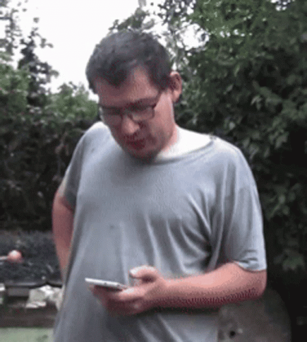 A Man Wearing Glasses And A Gray Shirt Is Looking At His Phone GIF