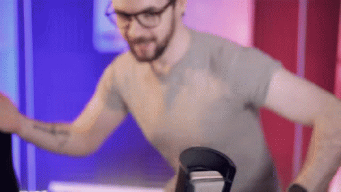 A Man Wearing Glasses And A Gray Shirt Is Standing In Front Of A Microphone . GIF