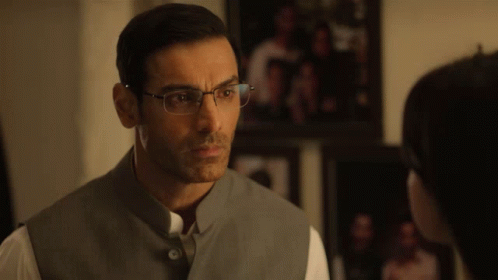A Man Wearing Glasses And A Gray Vest Looks At Another Man GIF