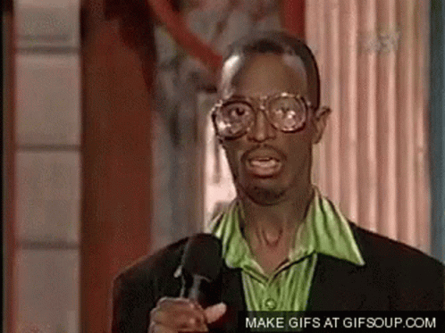 A Man Wearing Glasses And A Green Shirt Is Pointing At The Camera While Talking Into A Microphone . GIF