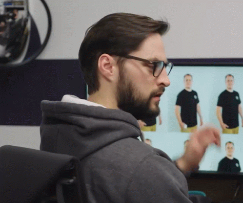 A Man Wearing Glasses And A Grey Hoodie Looks At A Monitor GIF