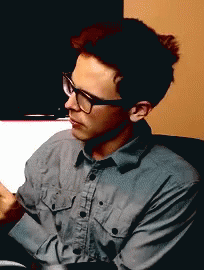 A Man Wearing Glasses And A Grey Shirt Is Sitting In A Chair GIF