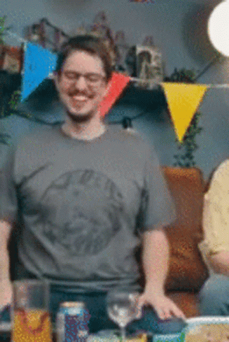 A Man Wearing Glasses And A Grey Shirt Is Standing In Front Of A Table GIF