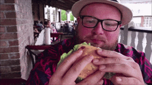 A Man Wearing Glasses And A Hat Is Eating A Burger GIF