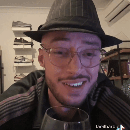 A Man Wearing Glasses And A Hat Is Holding A Glass Of Wine GIF