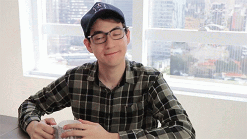 A Man Wearing Glasses And A Hat Is Sitting At A Table GIF