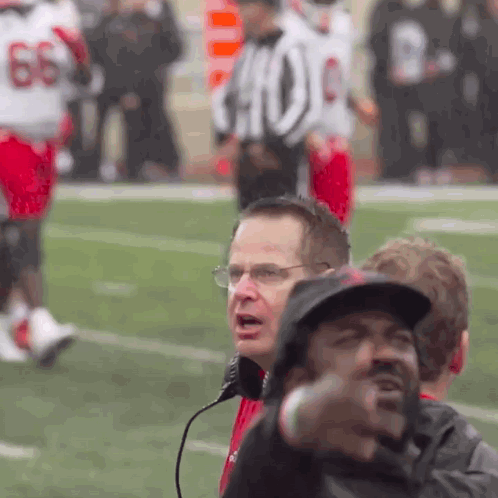 A Man Wearing Glasses And A Hat Stands On A Football Field In Front Of A Referee With The Number 66 On His Jersey GIF