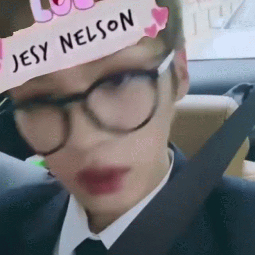 A Man Wearing Glasses And A Hat With The Name Jesy Nelson On It Is Sitting In A Car . GIF