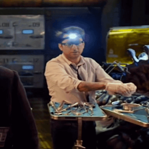 A Man Wearing Glasses And A Head Light Stands In Front Of A Table With Surgical Instruments On It GIF