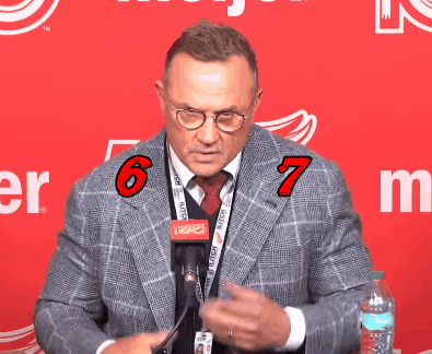 A Man Wearing Glasses And A Lanyard With The Number 6 And 7 On It Stands In Front Of A Red Background GIF