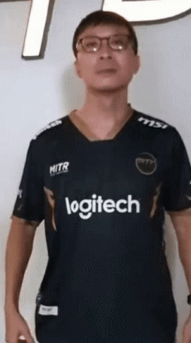 A Man Wearing Glasses And A Logitech Shirt Stands In Front Of A White Wall GIF