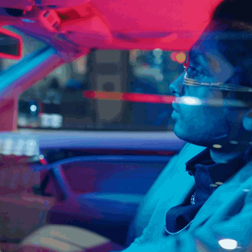 A Man Wearing Glasses And A Necklace Is Driving A Car At Night GIF