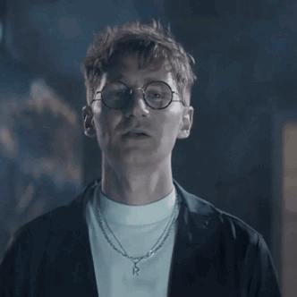 A Man Wearing Glasses And A Necklace Is Looking At The Camera In A Dark Room . GIF