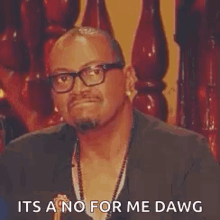 A Man Wearing Glasses And A Necklace Is Sitting In Front Of A Wooden Railing And Making A Funny Face . GIF