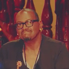 A Man Wearing Glasses And A Necklace Is Sitting In Front Of A Wooden Staircase . GIF