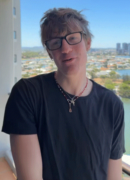 A Man Wearing Glasses And A Necklace Stands In Front Of A View Of A City GIF