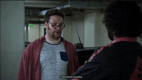 A Man Wearing Glasses And A Necklace Talks To Another Man In A Parking Garage GIF