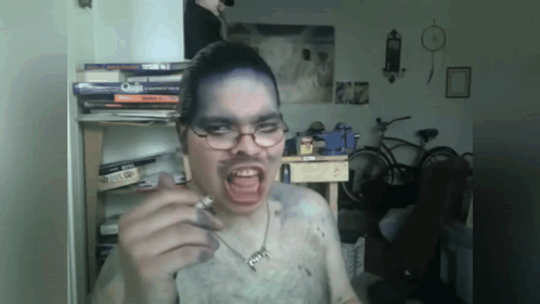A Man Wearing Glasses And A Necklace With The Letter G On It Is Smoking GIF