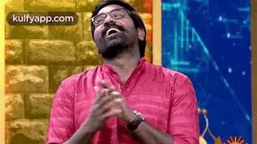 A Man Wearing Glasses And A Pink Shirt Is Clapping His Hands In Front Of A Brick Wall . GIF