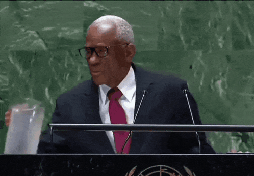 A Man Wearing Glasses And A Pink Tie Stands At A Podium GIF