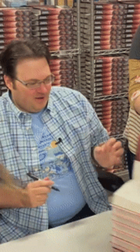 A Man Wearing Glasses And A Plaid Shirt Is Signing Books GIF