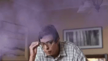 A Man Wearing Glasses And A Plaid Shirt Is Sitting In A Room With Purple Smoke Coming From His Head . GIF