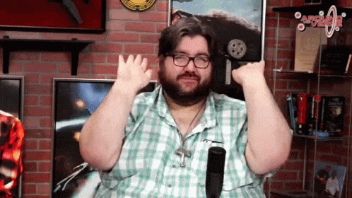 A Man Wearing Glasses And A Plaid Shirt Is Sitting In Front Of A Brick Wall With A Sign That Says Around The World GIF