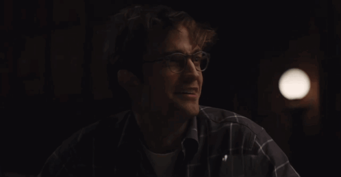 A Man Wearing Glasses And A Plaid Shirt Smiles In The Dark GIF