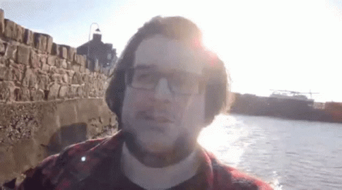 A Man Wearing Glasses And A Plaid Shirt Stands In Front Of A Body Of Water GIF
