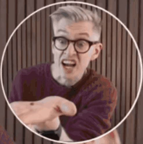 A Man Wearing Glasses And A Purple Shirt Is Pointing At The Camera . GIF