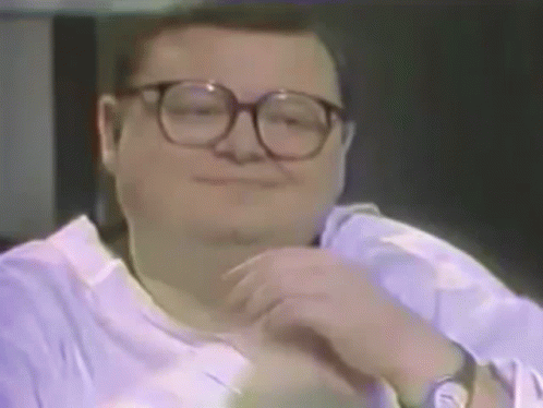 A Man Wearing Glasses And A Purple Shirt Is Smiling And Looking At The Camera . GIF