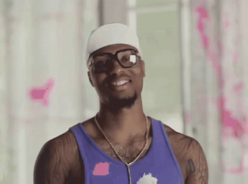 a man wearing glasses and a purple tank top smiles for the camera