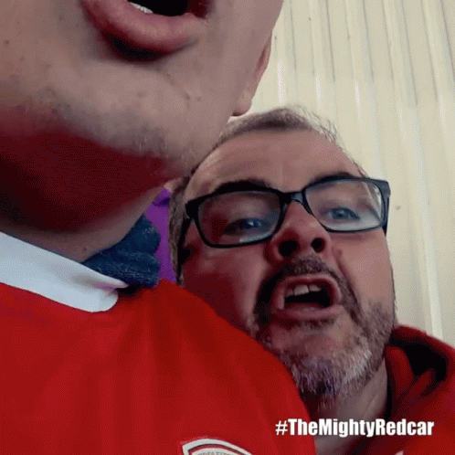 a man wearing glasses and a red shirt has #themightyredcar written on the bottom